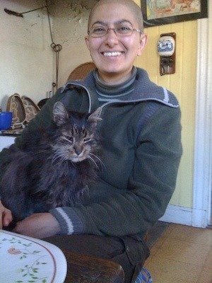 Leende person med glasögon håller en långhårig grå katt i famnen i ett gårdskök