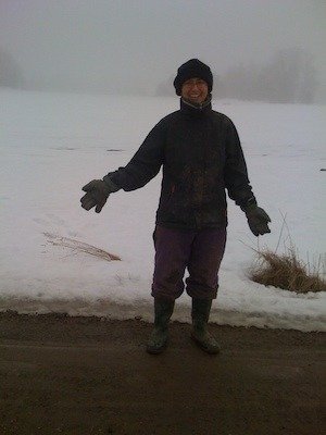 Person i mössa, arbetsjacka och gummistövlar står på en snöig väg vid ett åkerfält på Ramsjö gård
