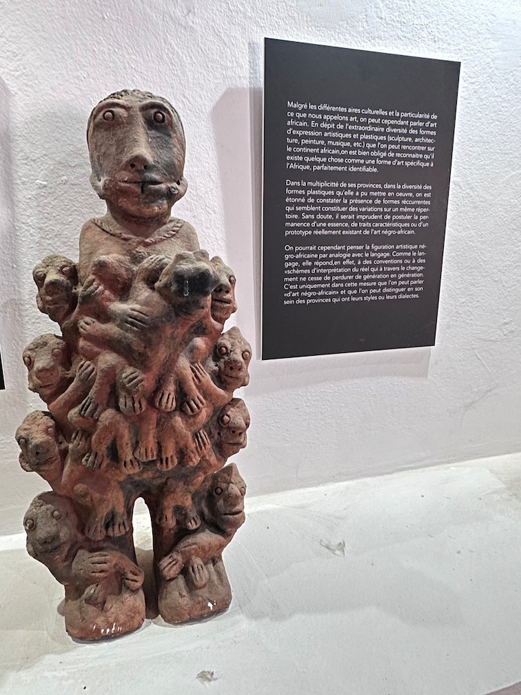 A clay sculpture with many small faces stands next to a text panel.
