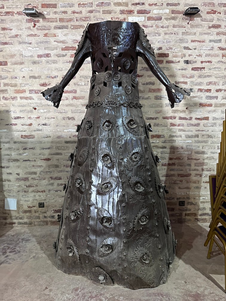 A metal sculpture shaped like a long dress stands in front of a brick wall.