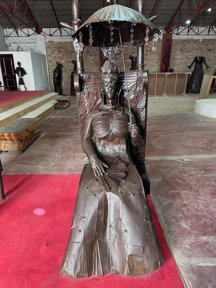 A metal sculpture of a seated woman with an umbrella stands in a gallery.