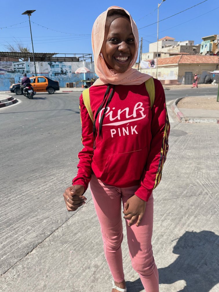 A smiling girl in a red hoodie stands on a street in Saint-Louis.