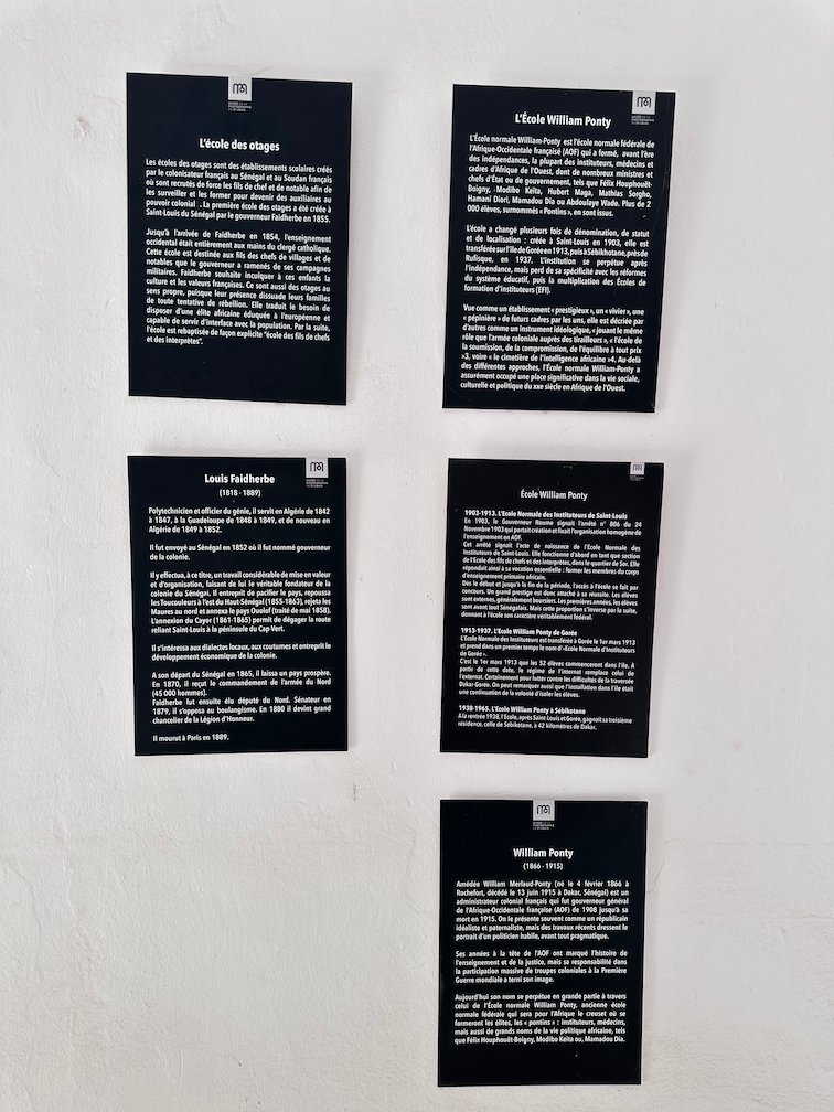 Six black text panels hang on a white wall in a museum in Saint-Louis.