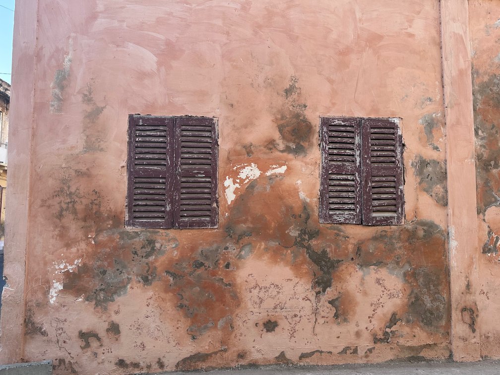 Two wooden shutters sit in a worn pink wall in Saint-Louis.