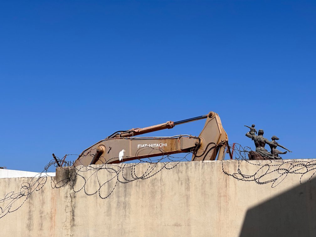 An excavator stands behind a wall with razor wire in Saly.