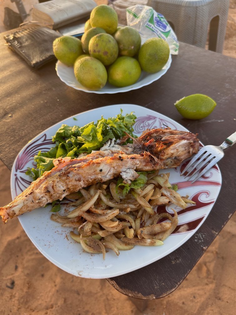 A plate of grilled fish with onions stands on a table on the sand.