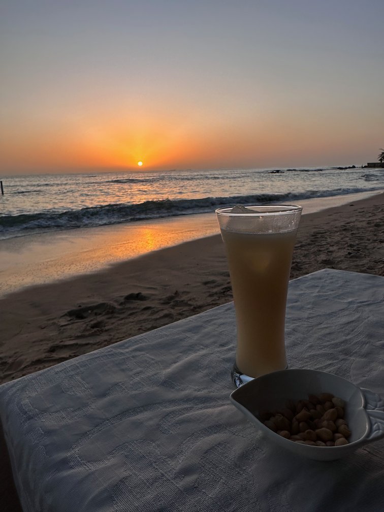 A drink and a bowl of peanuts stand on a table at sunset on the beach in Saly.