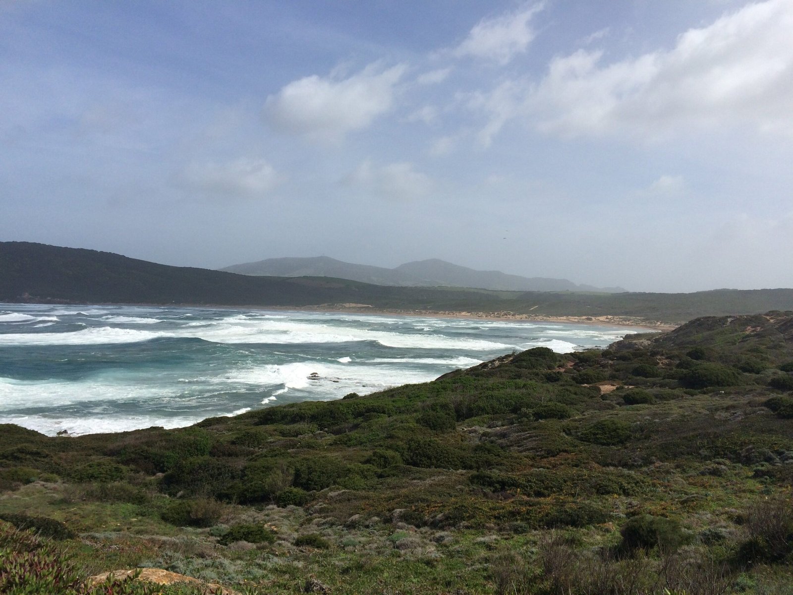 White waves roll onto a long beach below green hills on the coast of Sardinia.