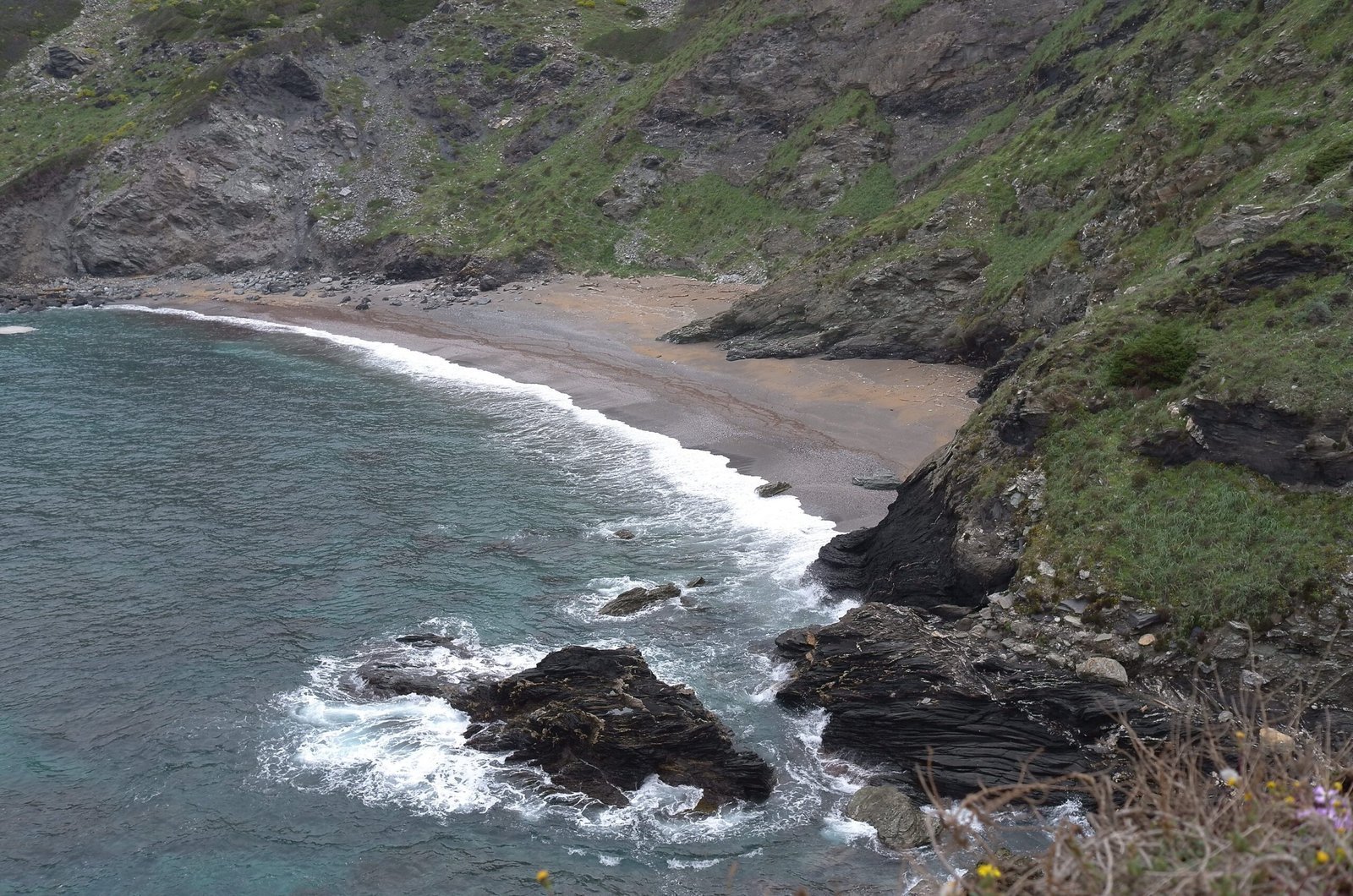 A curved beach and rocky shore lie below green slopes on the coast of Sardinia.