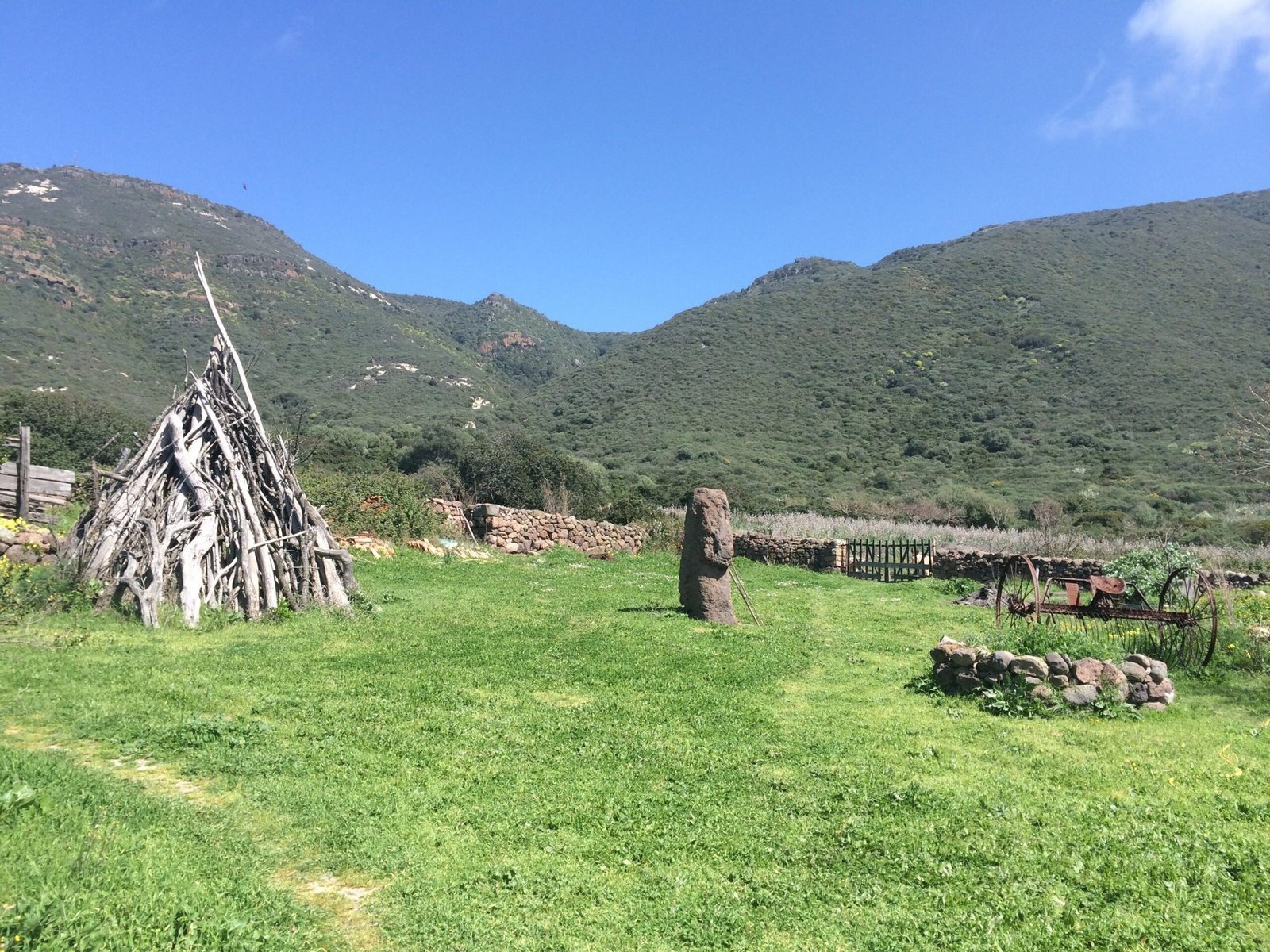 A wooden teepee and a stone stand in a green garden below hills in Sardinia.