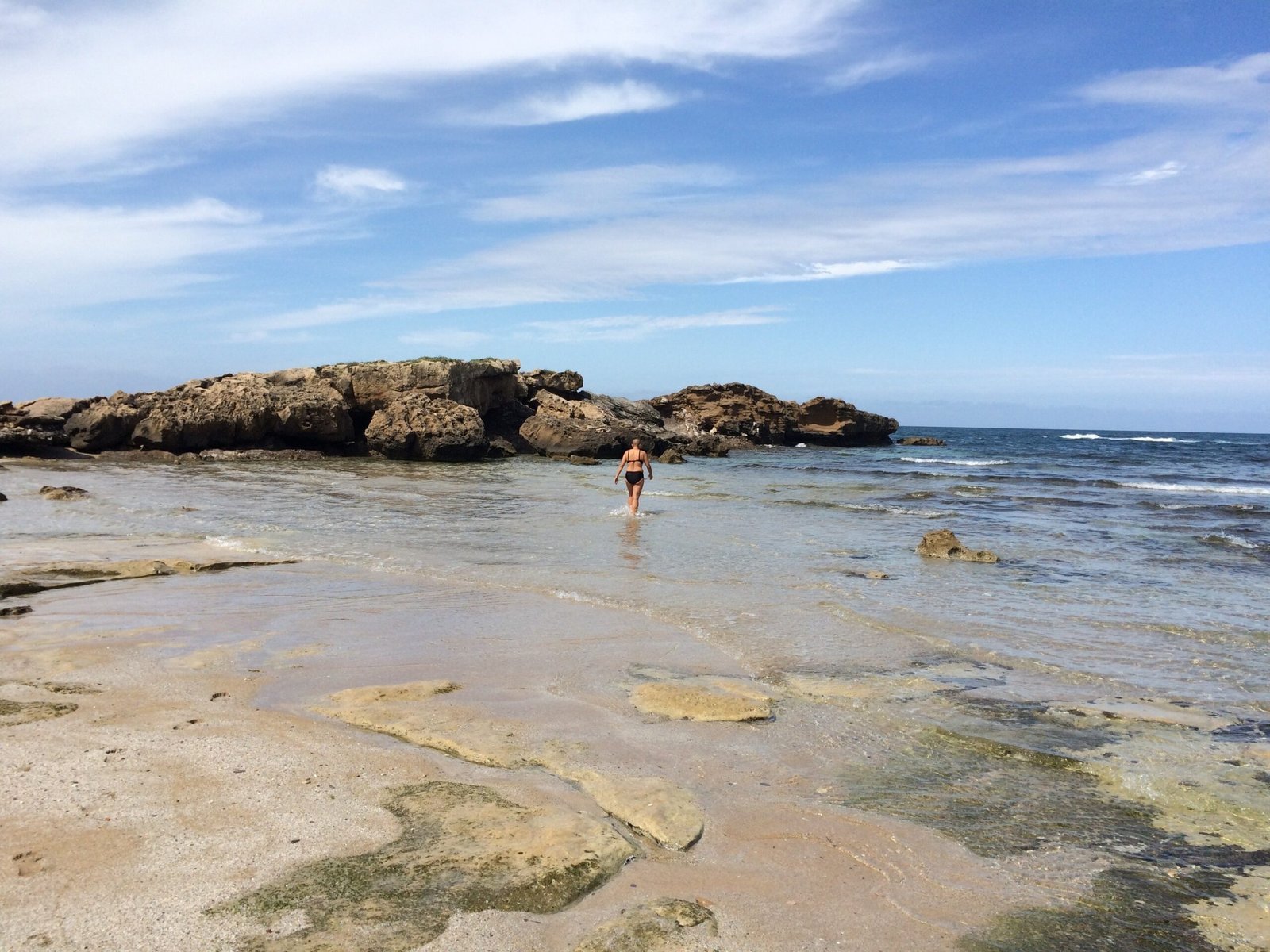 A person wades in shallow water near flat rocks on the coast of Sardinia.