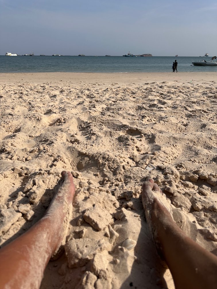Bare feet rest in the sand in front of the calm sea.