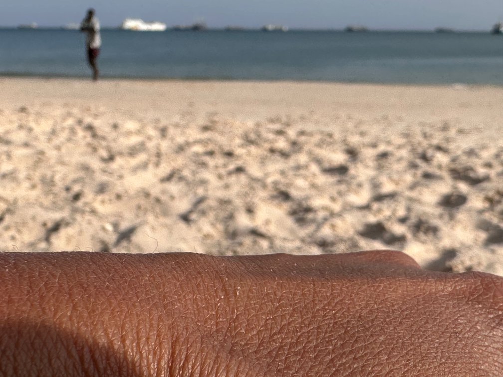 A hand rests on the sand while a person stands by the sea.