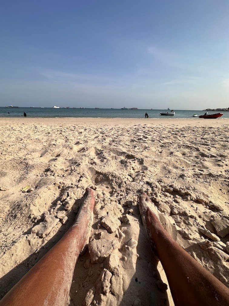 Legs rest on the sand of a beach with boats in the sea.