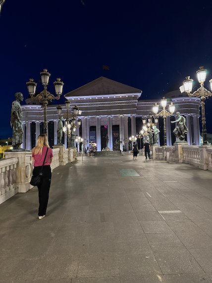 A bridge with lamps and statues leads to the Archaeological Museum at night in Skopje.