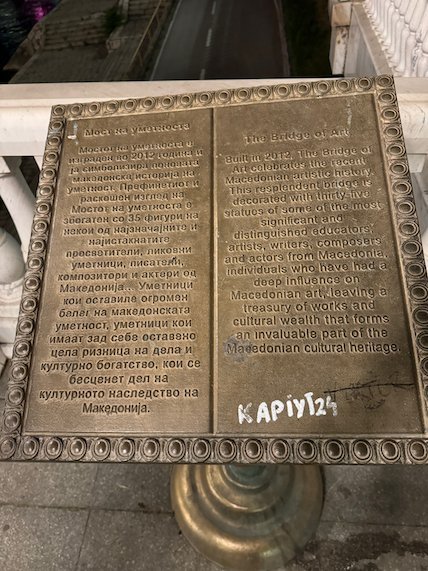 A bronze plaque with text in Macedonian and English stands on a bridge in Skopje.