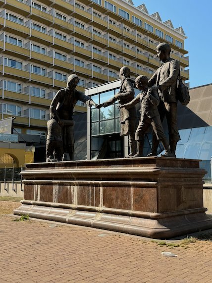 A bronze statue of a family stands on a stone base in front of a hotel in Skopje.