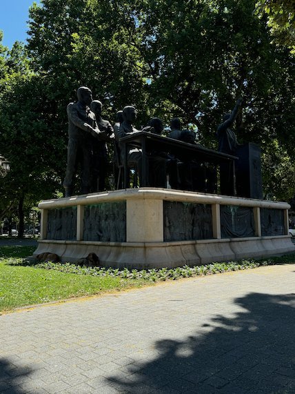 Bronze figures sit around a table on a stone base in a park in Skopje.
