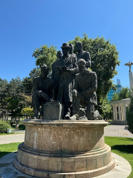 A bronze group of men stands on a round stone base in a park in Skopje.