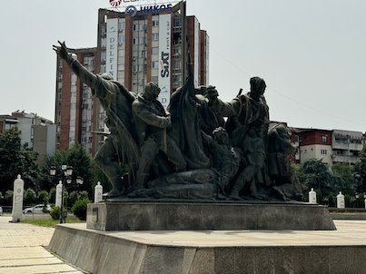 A bronze monument of figures with raised arms stands in front of tall buildings in Skopje.