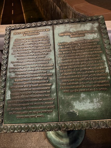 A bronze plaque with two text panels stands on a bridge in Skopje.