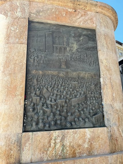 A bronze relief panel shows a crowd and buildings on a monument base in Skopje.