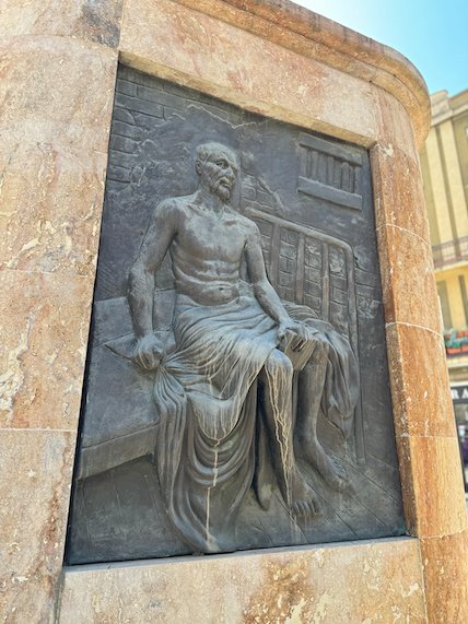 A bronze relief shows a seated man on a stone monument base in Skopje.