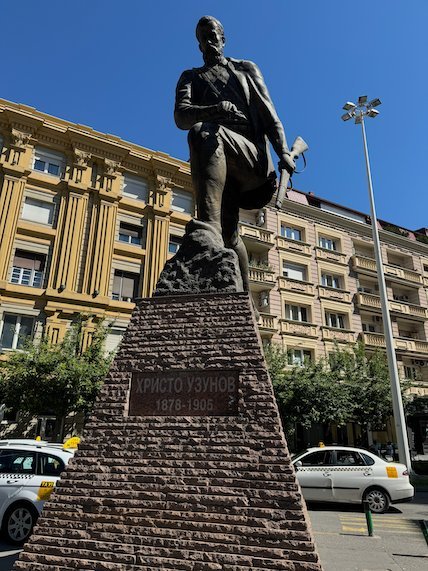 A bronze statue of a man stands on a red stone base in central Skopje.
