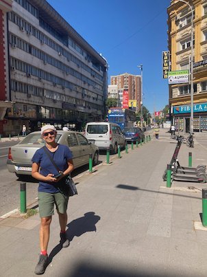 A traveler walks along a sunny city street with parked cars in Skopje.