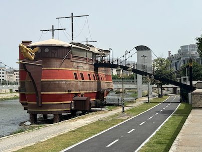 A large wooden galleon ship stands on the bank of the Vardar River beside a bike path in Skopje.