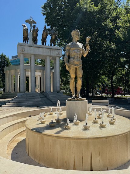 A gold statue stands on a fountain in front of a colonnade with statues in Skopje.