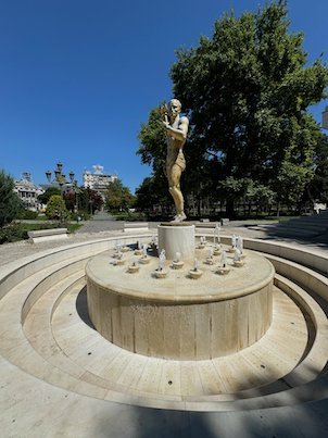 A gold statue stands on a round fountain in a park in Skopje.