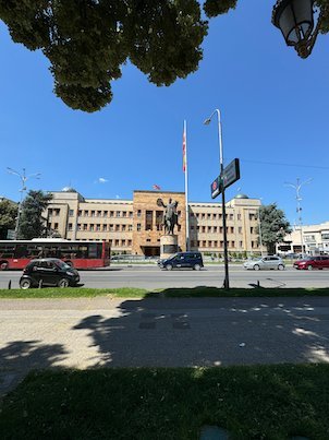 A large government building and an equestrian statue stand across a busy street in Skopje.