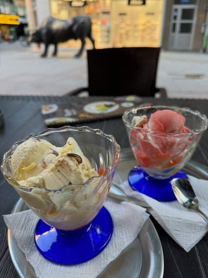 Two cups of ice cream sit on a cafe table with a bull statue behind in Skopje.