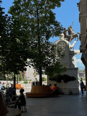 A white monument with a horse on top stands behind trees and cafe seats in Skopje.