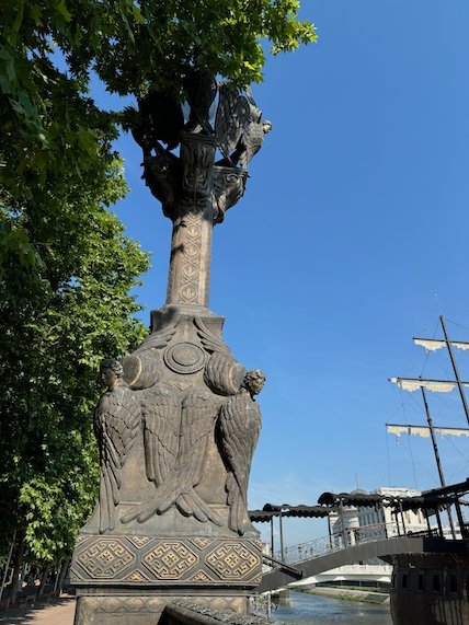 An ornate column with carved eagles stands near the river, with a galleon behind, in Skopje.