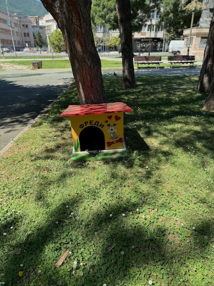 A small painted dog house stands under a tree in a park in Skopje.
