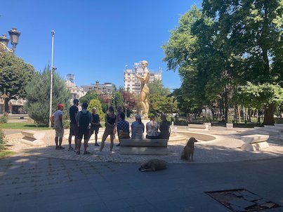 A group of people stands near a gold statue in a park with a dog lying nearby in Skopje.