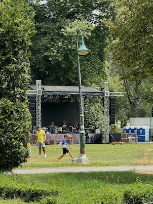 An outdoor stage stands on a lawn with people walking past in a park in Skopje.