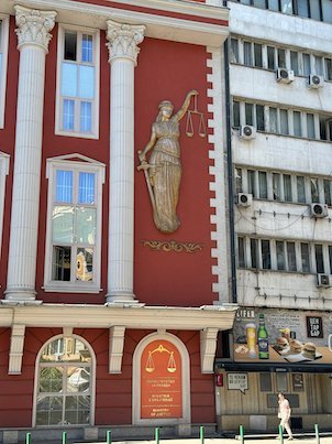 A gold Lady Justice figure decorates a red building facade with white columns in Skopje.