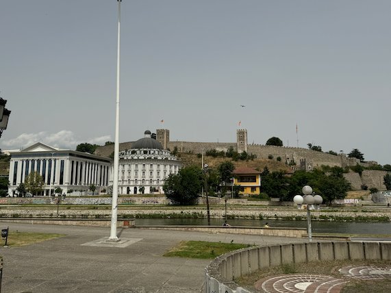 A tall flagpole stands on a riverside square with Kale Fortress on the hill in Skopje.