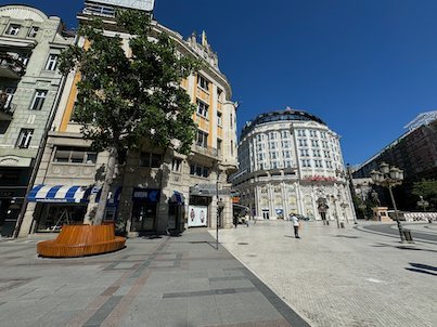 A round wooden bench stands on a square in front of old buildings in Skopje.