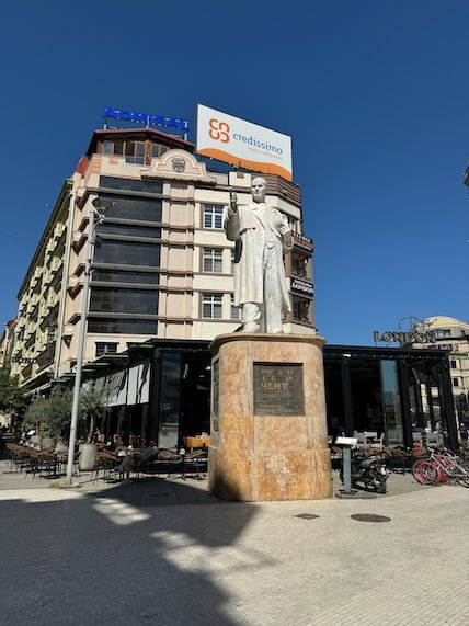 A white statue stands on a tall pedestal at a street corner in Skopje.