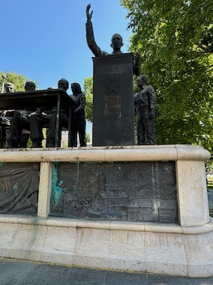 A bronze statue of a man with a raised arm stands above a relief base in Skopje.