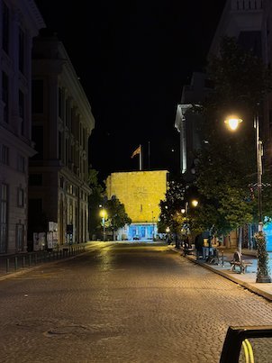 A street at night leads to a brightly lit yellow building in Skopje.