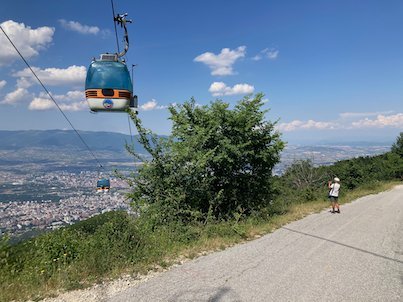 A cable car cabin passes above a mountain road with a view over Skopje from Mount Vodno.