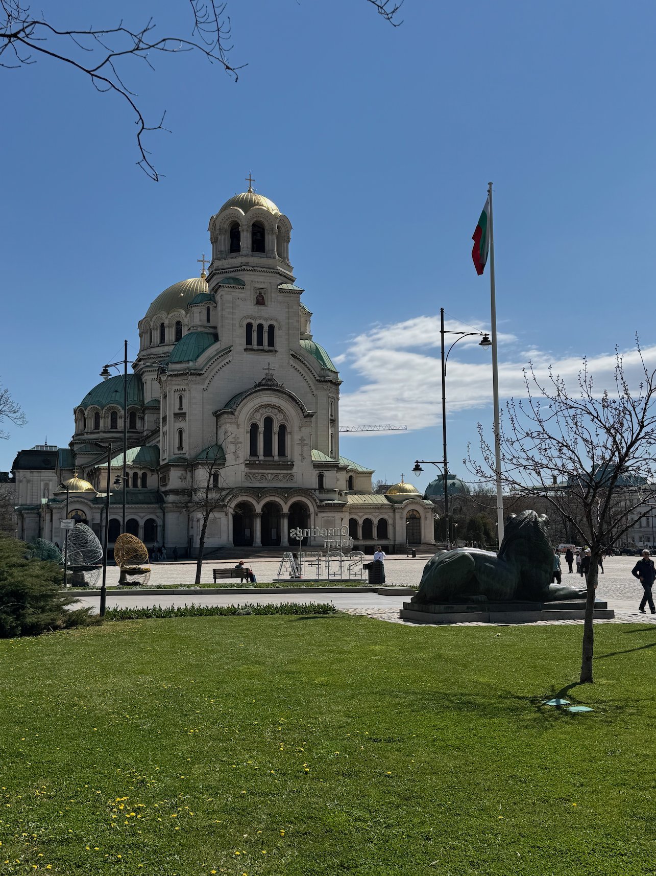 Alexander Nevsky Cathedral stands behind a green lawn and a Bulgarian flag in Sofia.