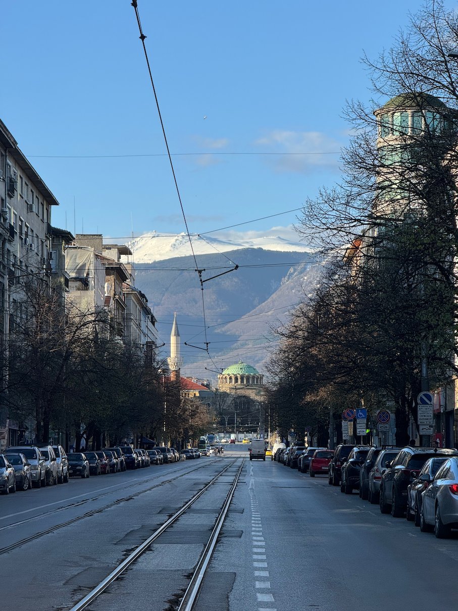 A wide boulevard with tram tracks leads to a golden dome and snowy Vitosha Mountain in Sofia.