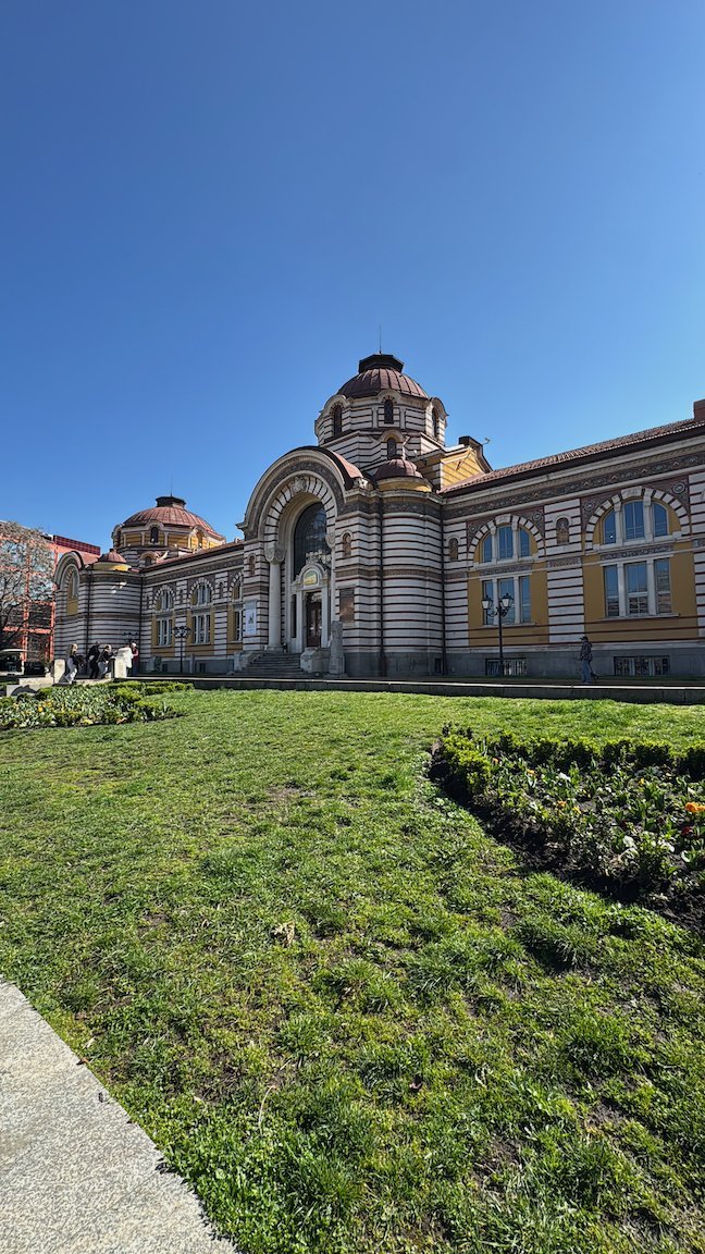 The yellow and red Central Mineral Baths building stands behind a green lawn in Sofia.