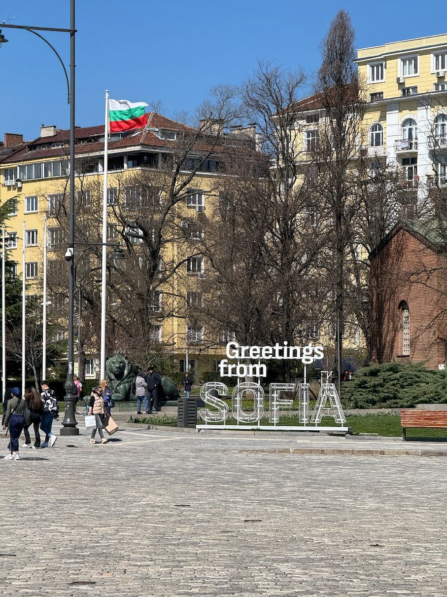 A Greetings from Sofia sign stands on a square with a Bulgarian flag behind it in Sofia.